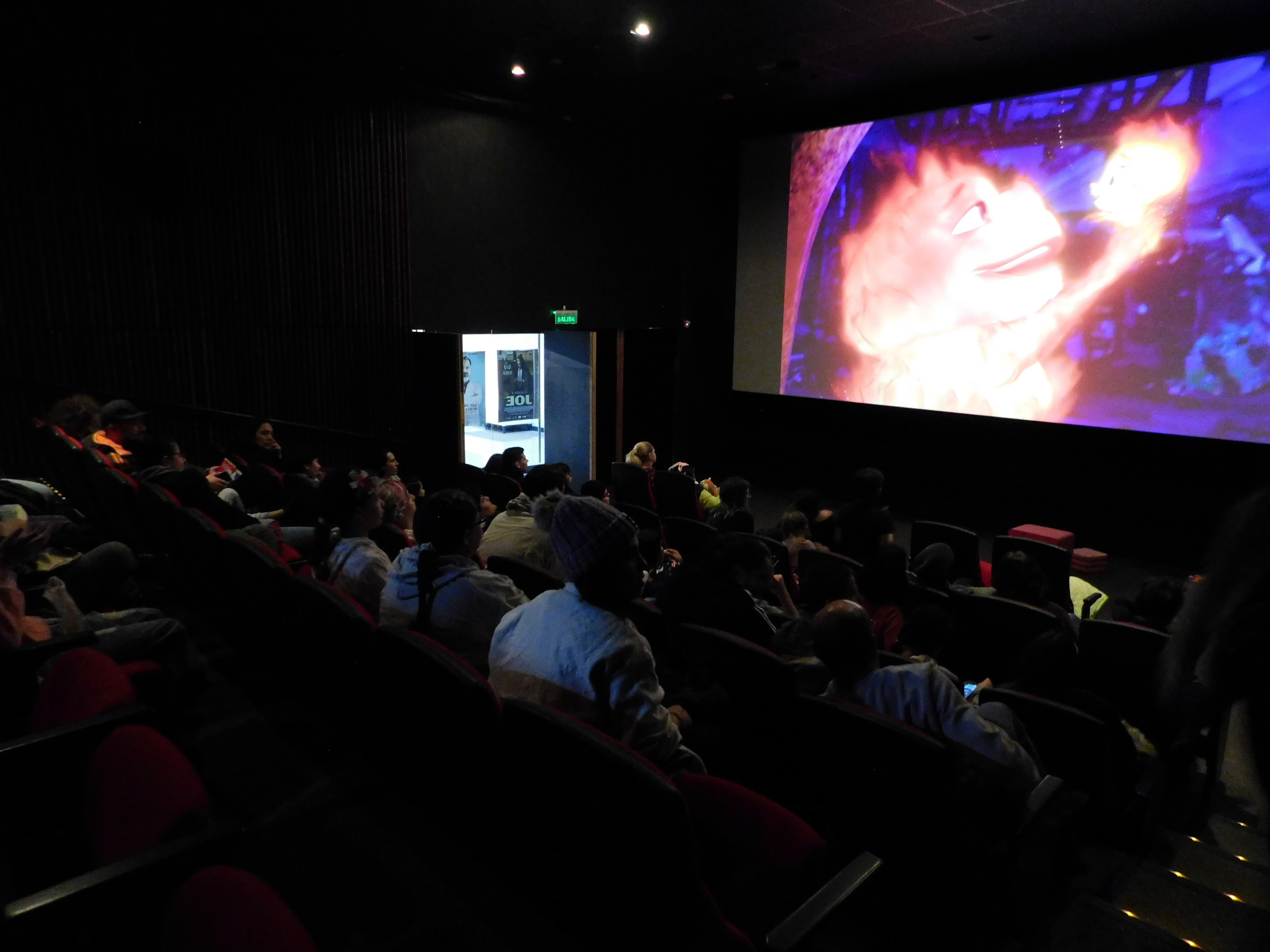 Fotografía tomada en lateral dentro de la sala de cine donde se observa a los asistentes de espaldas y de frente la pantalla de cine
