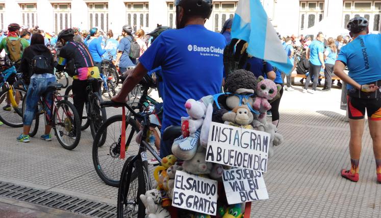 Fotografia de una persona ciclista de espaldas con distintos carteles y peluches colocados en la bicicleta