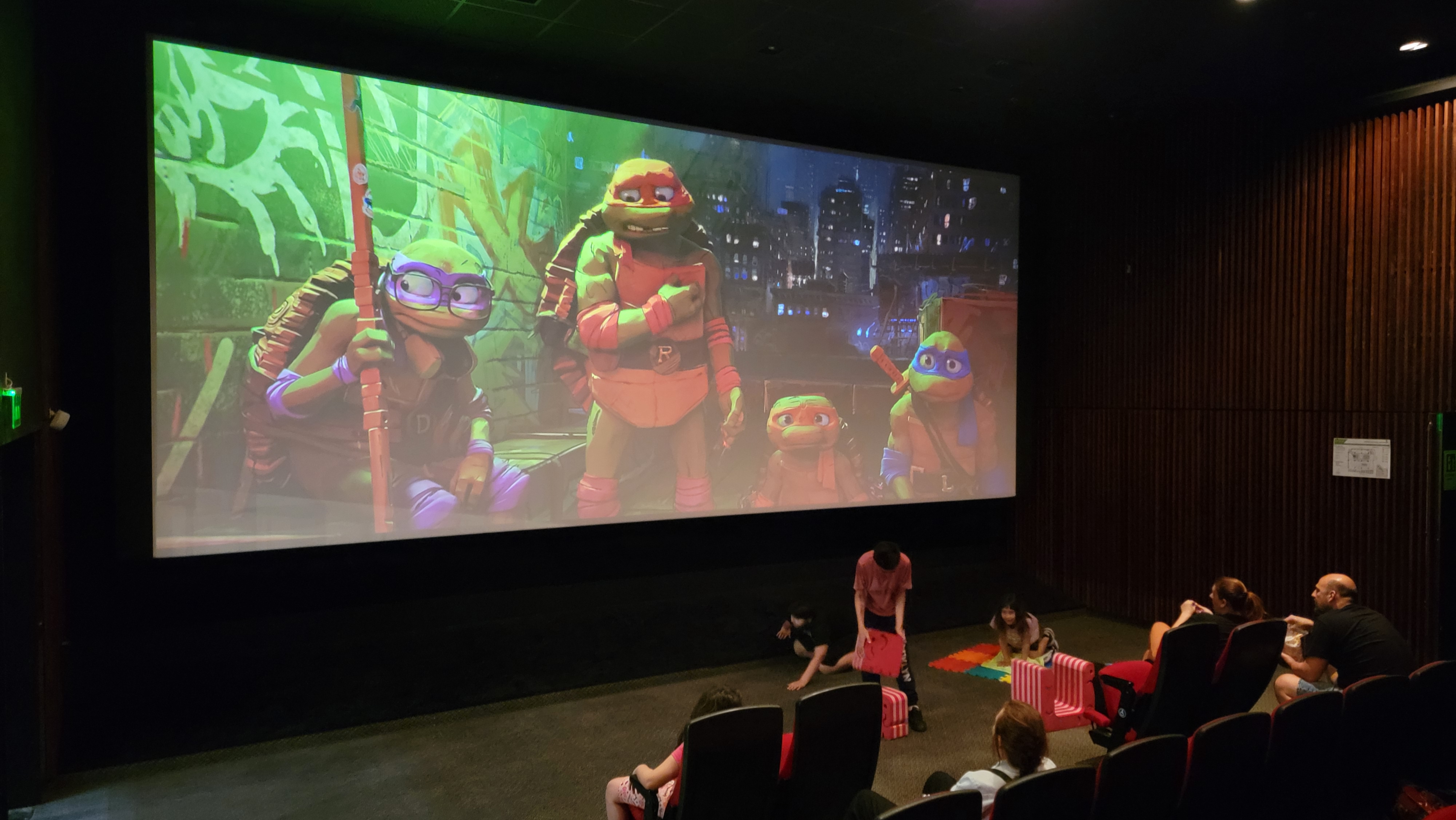 Fotografia tomada en lateral y dentro de la sala donde se observa la pantalla con una escena con los personajes. Parte de los chicos jugando con la sala de apoyo