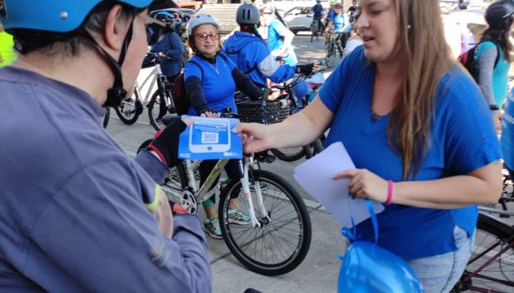 Imagen 4: Del lado derecho la imagen de una mujer vestida de color azul con un globo azul en la mano entregando un folleto a un hombre en bicicleta y con un casco que se encuentra del lado izquierdo de la imagen.
